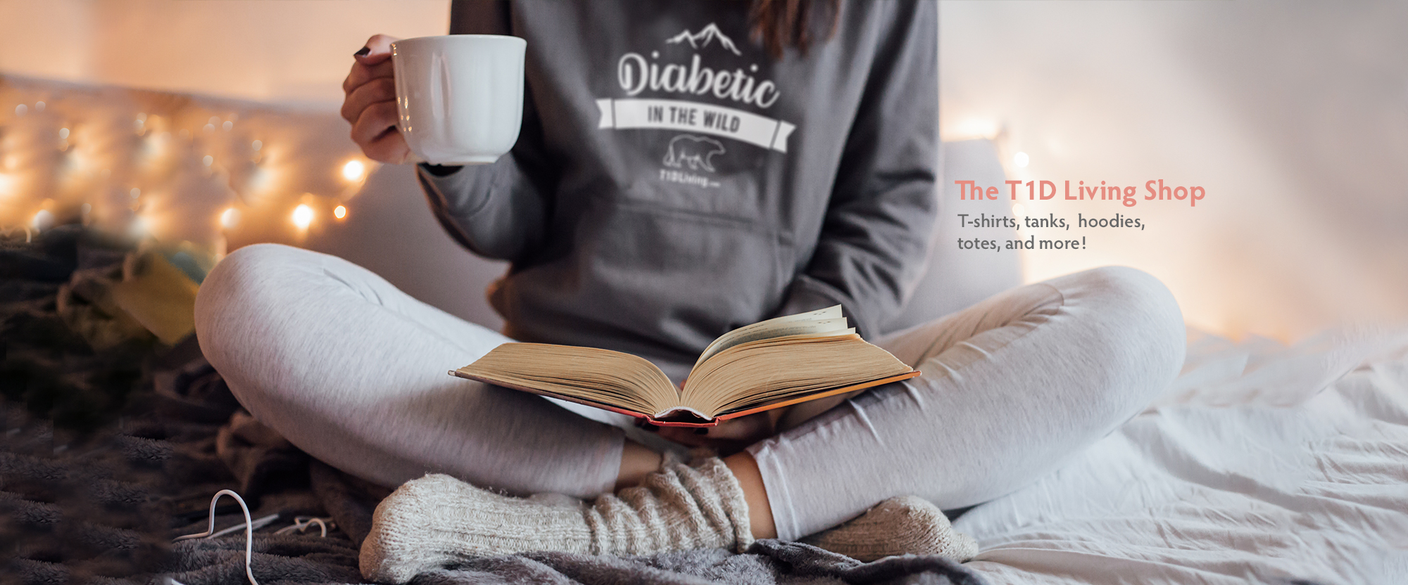 Woman in cozy sweatpants and T1D branded hoodie on bed reading a book and drinking tea